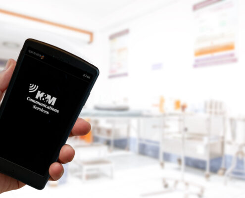 K&M Communication Services logo on screen of Black Spectralink Pivot 8744 smartphone on a medical blur background